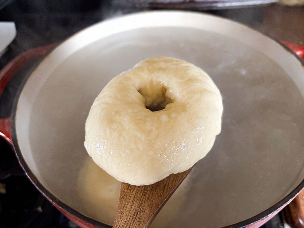 Sourdough bagel being lifted from a pot of boiling baking soda water on a wooden spoon.