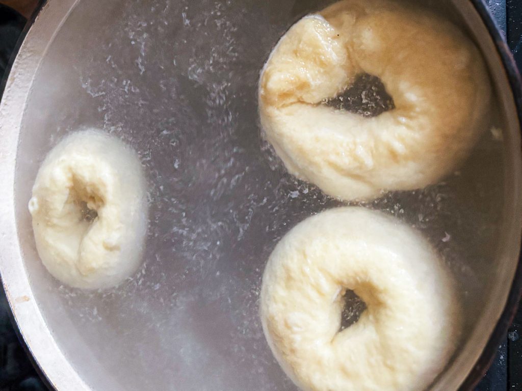 Three sourdough bagels boiling in a pot of bubbling baking soda water.