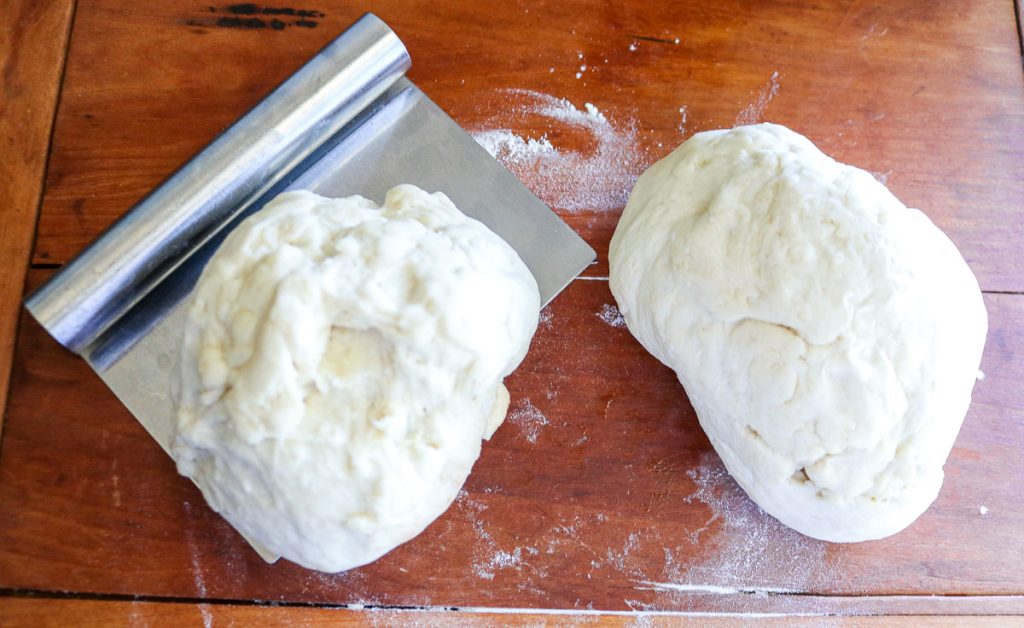 two halves of bread dough with a bench scraper on a wooden cutting board