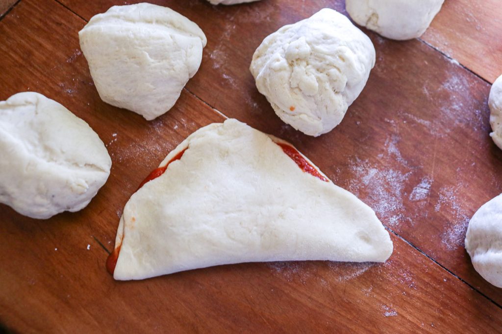 unbaked circles of dough on a wooden board with one piece spread with marinara and rolled up