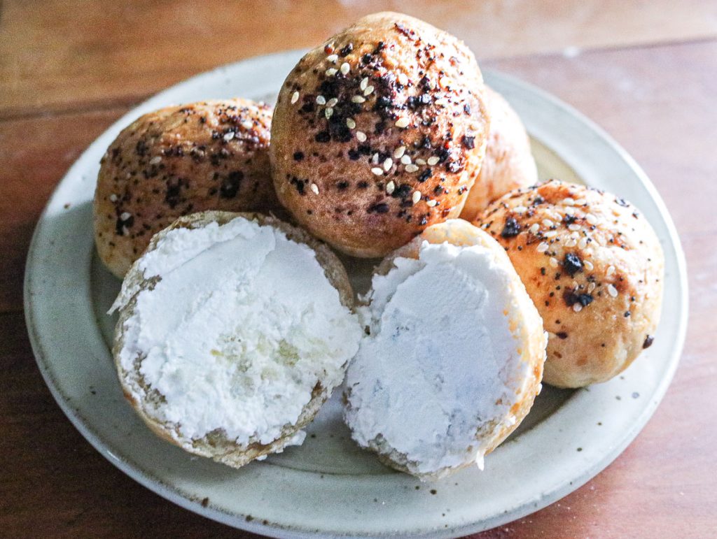 stack of miniature bagels on a plate with one cut in half and spread with cream cheese