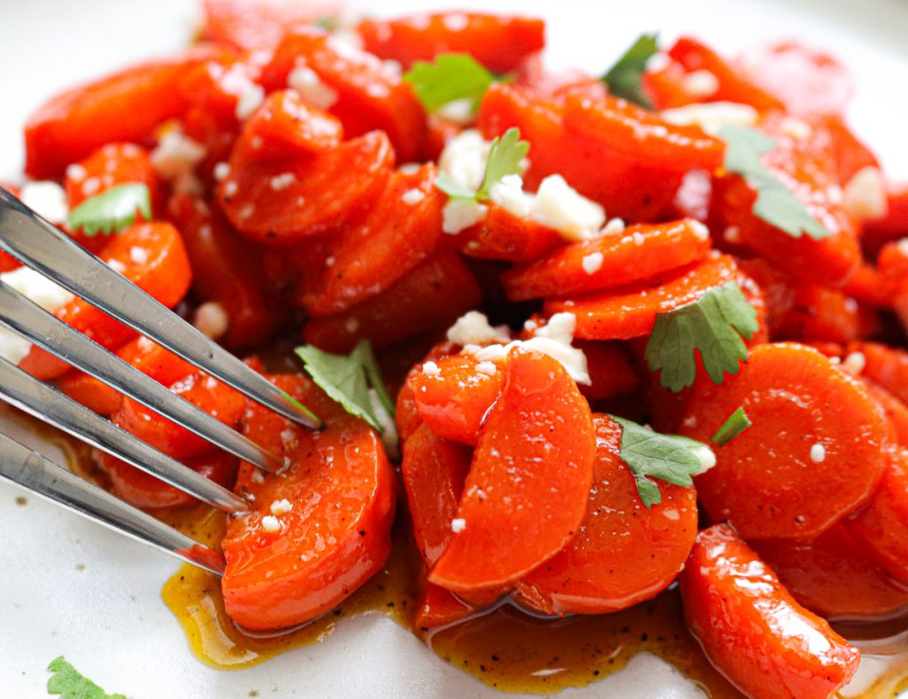 a fork piercing a glazed carrots on a white plate with feta and herbs