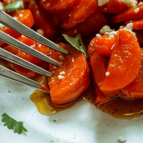 Close-up fork taking a bite of hot honey glazed carrots topped with feta and fresh herbs
