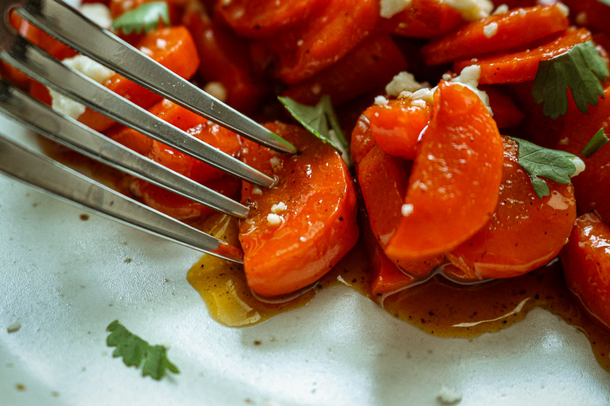 Close-up fork taking a bite of hot honey glazed carrots topped with feta and fresh herbs
