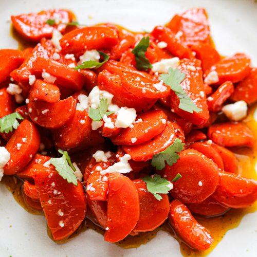 a white plate of sliced glazed carrots with feta and herbs