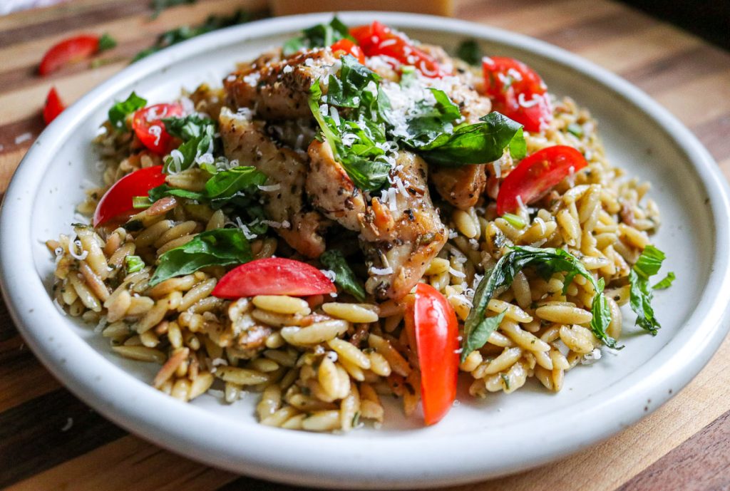 white plate of pesto orzo with chicken and tomatoes on a wooden counter top with parmesan cheese and grape tomatoes in the background