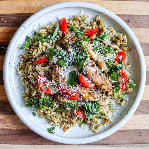 white plate of pesto orzo with chicken and tomatoes on a wooden counter top with parmesan cheese and grape tomatoes in the background