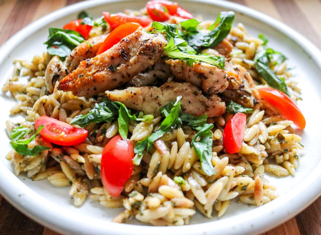 white plate of pesto orzo with chicken and tomatoes on a wooden counter top