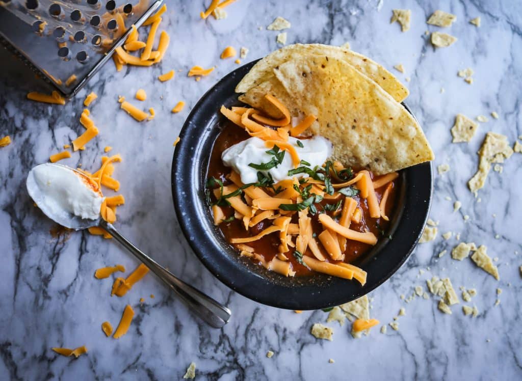 taco soup in a black bowl with tortillas, shredded cheese and sour cream on a white marble counter top with cheese and chips on it