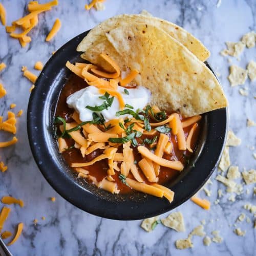 taco soup in a black bowl with tortillas, shredded cheese and sour cream on a white marble counter top with cheese and chips on it