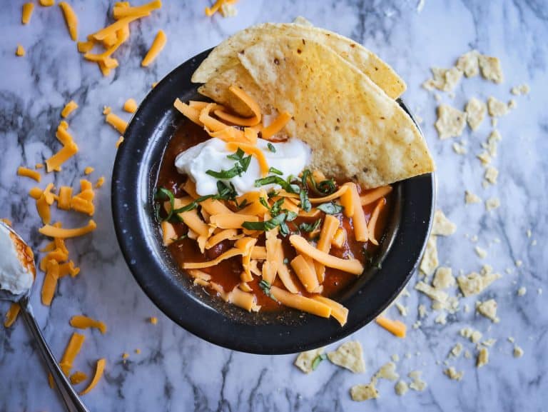 taco soup in a black bowl with tortillas, shredded cheese and sour cream on a white marble counter top with cheese and chips on it