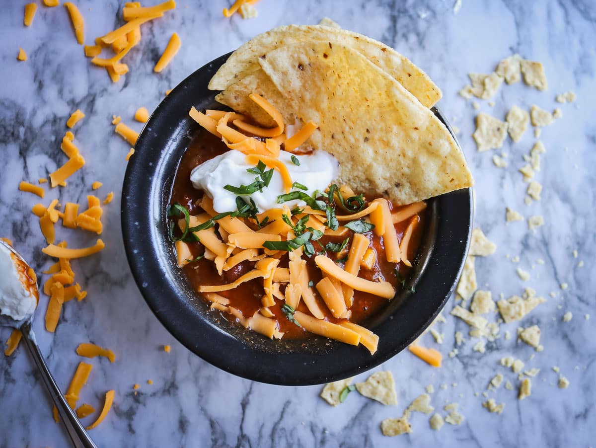 taco soup in a black bowl with tortillas, shredded cheese and sour cream on a white marble counter top with cheese and chips on it