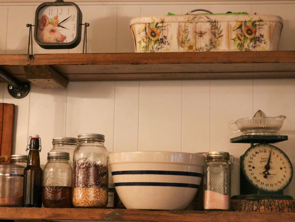 From scratch pantry shelves with glass jars of grains, cornmeal, and pantry staples alongside a mixing bowl and vintage kitchen scale.