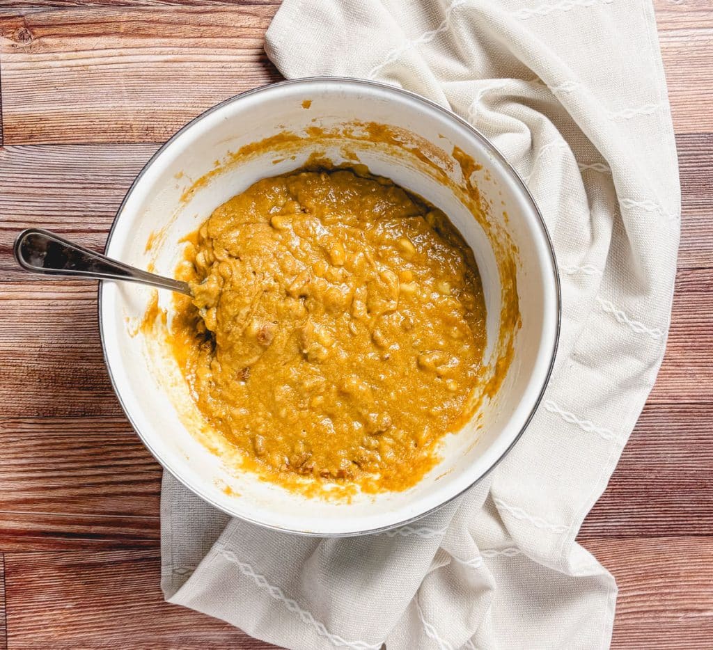 Banana bread batter with pecans in a white mixing bowl on a wooden surface next to a cream-colored towel.