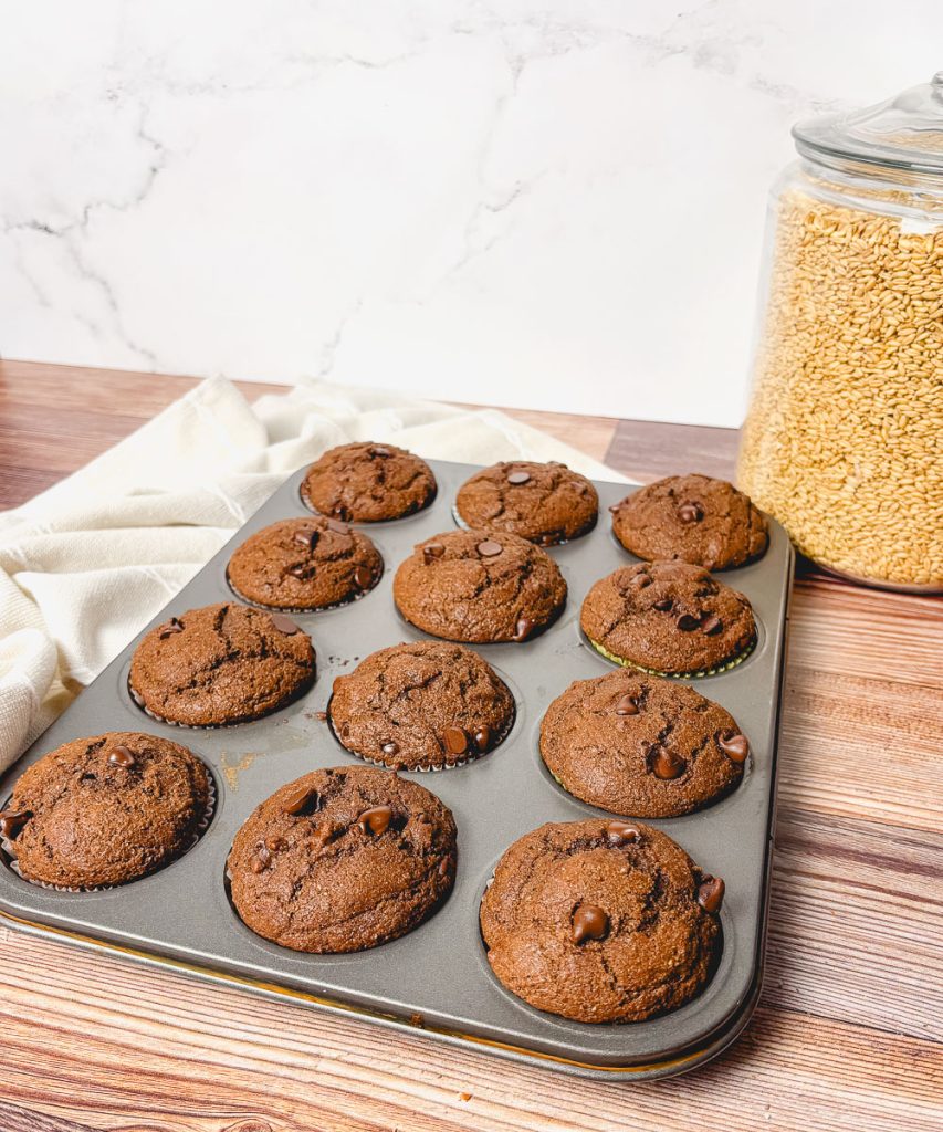side angle of a full tray of baked double chocolate chip muffins with a glass container of wheat berries in the background