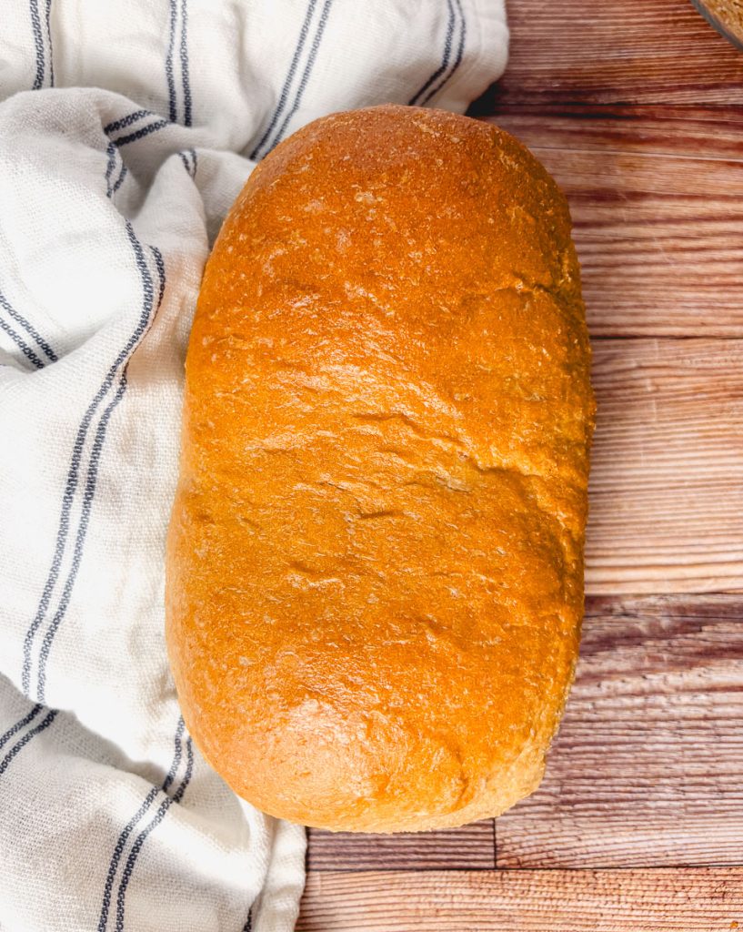 Whole loaf of honey wheat bread made with fresh-milled flour on a wooden board next to a striped kitchen towel.
