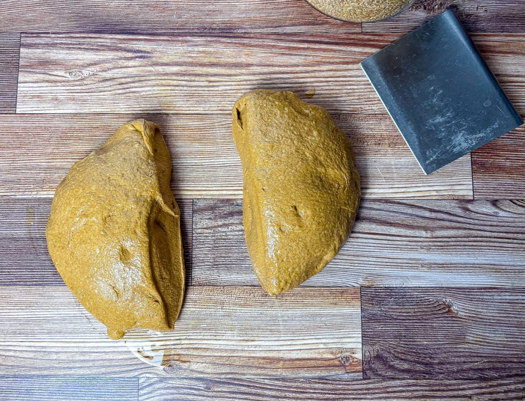 brown bread dough split into two pieces with a bench scraper