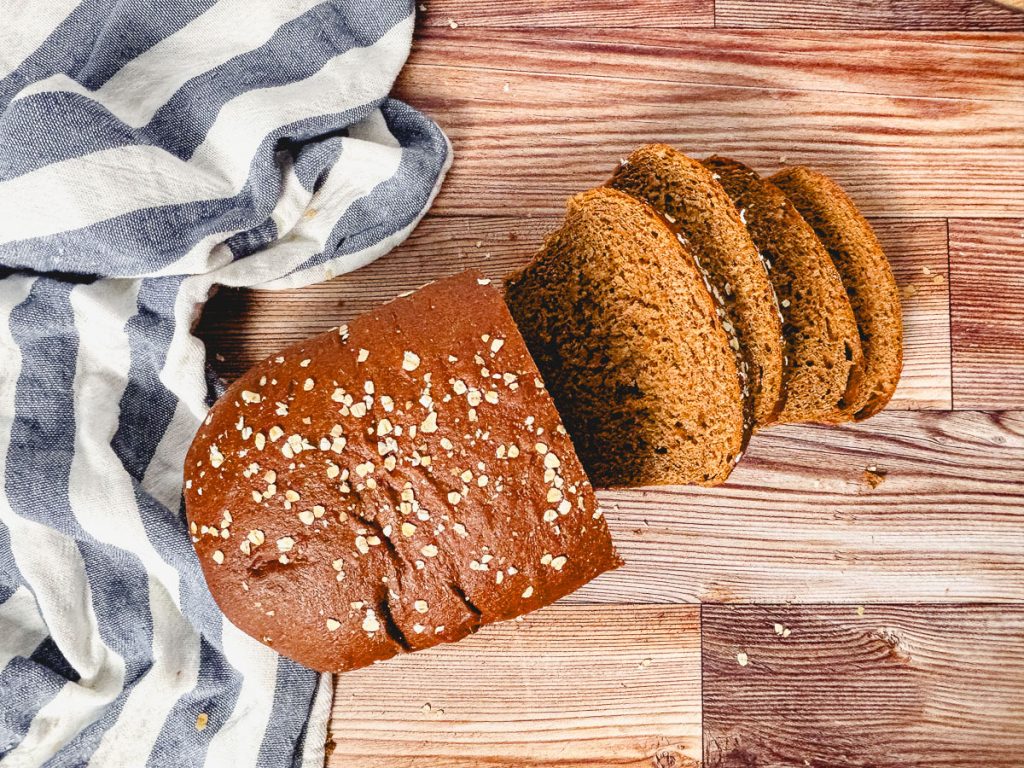 a partially sliced brown bread loaf on a wooden counter with a striped blue and white tea towel
