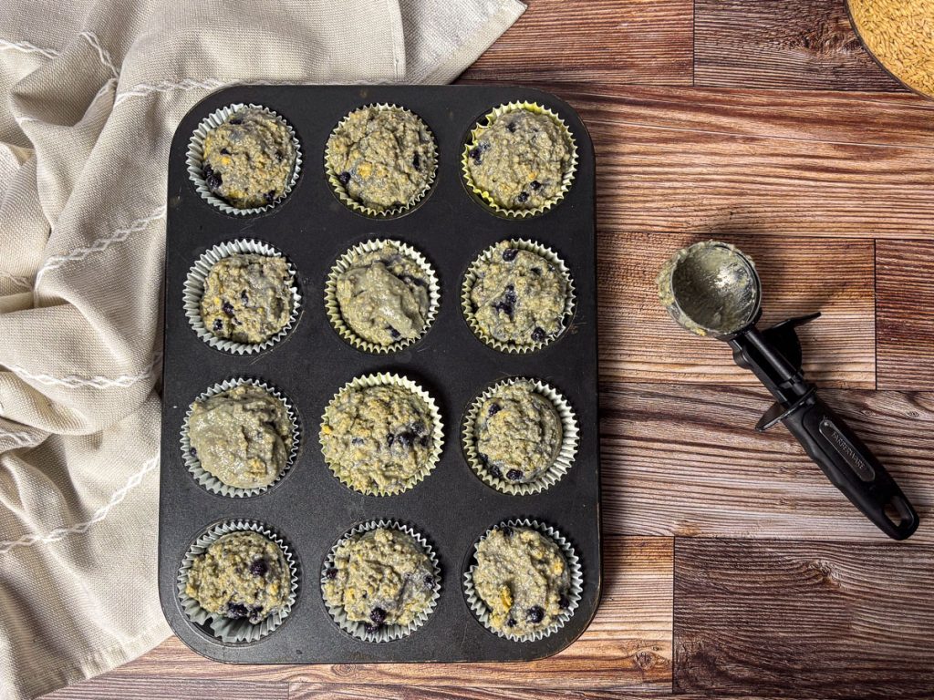 full tray of unbaked blueberry muffins with a cookie scoop and tea towel