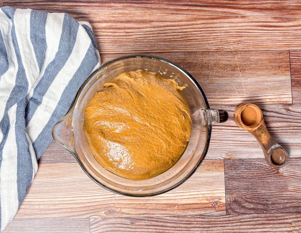 unproofed brown bread dough in a glass bowl with a blue and white striped tea towel and measuring spoon