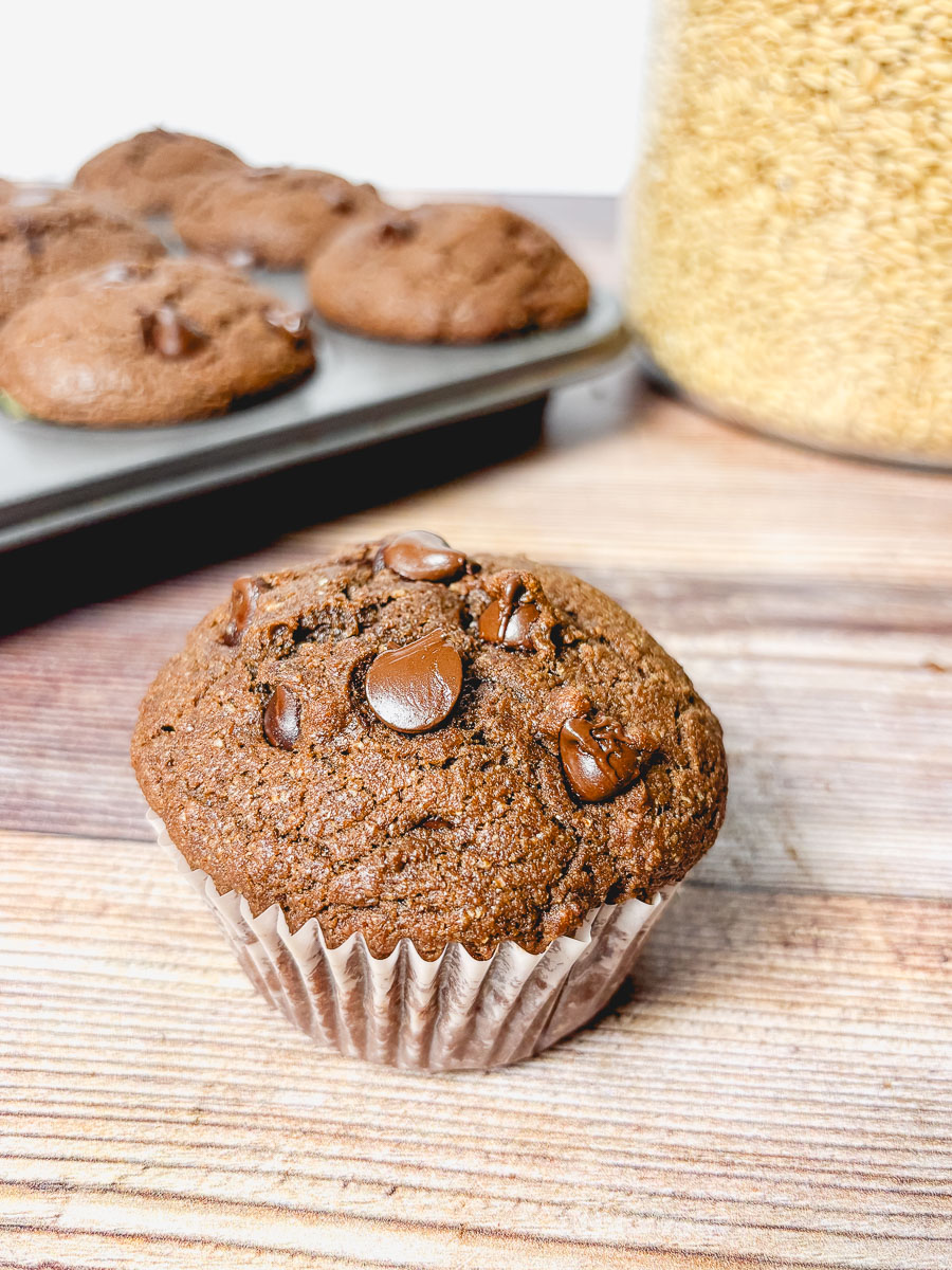 upclose double chocolate chip muffin in front of a full tray of muffins and a glass container of wheat berries