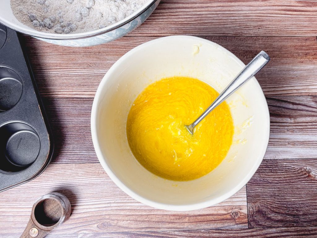 mixed wet ingredients for blueberry muffins with measuring spoon, muffin tray and dry ingredients in a bowl in the background