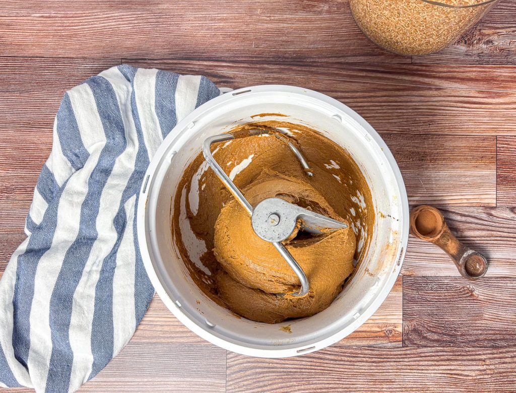 a zoomed out view of kneaded brown bread in a bosch mixer bowl with a blue and white tea towel, measuring spoon and wheat grain in the background